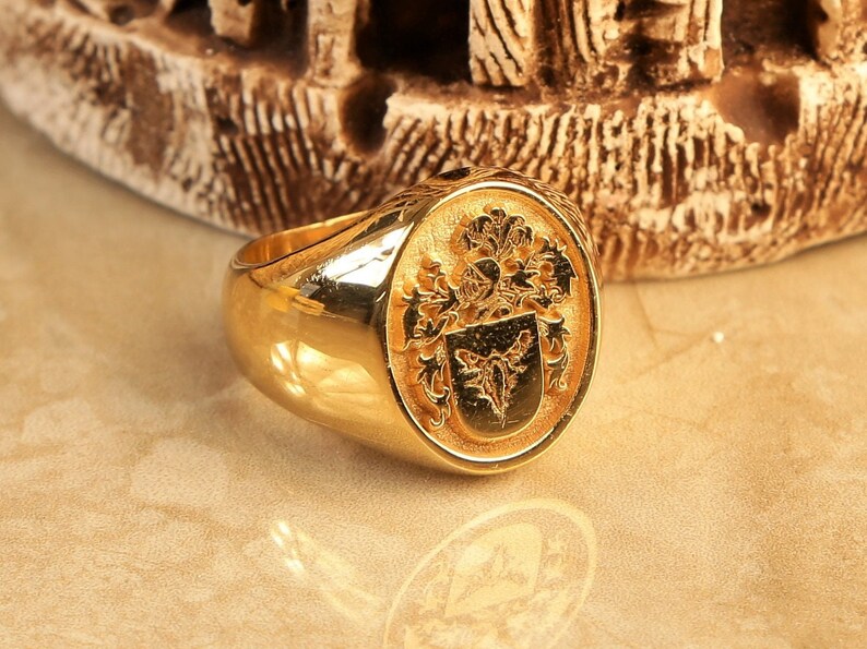 May include: A gold signet ring featuring a detailed crest design. The crest includes a shield with a stylised animal and floral embellishments.