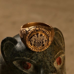 May include: A gold ring featuring a detailed crest with two lions and a shield with stripes. The ring is set on a dark green stone surface.