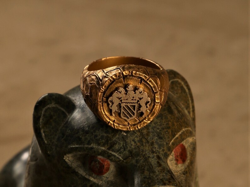 May include: A gold ring featuring a detailed crest with two lions and a shield with stripes. The ring is set on a dark green stone surface.