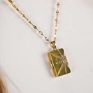 Puede incluir: Collar con colgante rectangular dorado con diseño de sol. El colgante está adornado con pequeños cristales brillantes. La cadena del collar es una delicada cadena de eslabones dorados.
