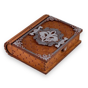 Puede incluir: Una caja de madera con diseño de libro. La caja es marrón con un patrón de diamantes y detalles plateados. Un diseño de flor de lis plateada está centrado en la tapa. Un emblema de corona es visible en el lateral.