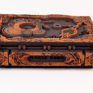 May include: A wooden box with a dark brown finish and intricate carvings. The top of the box features a steampunk design with gears, a dragonfly, and a clock face. The front of the box has the words "MAGIC BOOK" engraved on it.