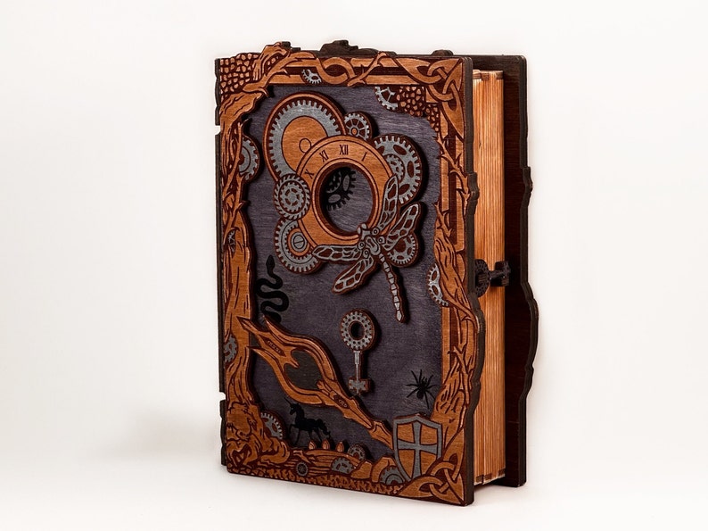 May include: A wooden book with a dark gray cover and intricate carvings. The cover features a dragonfly, gears, and other steampunk-inspired designs. The book is closed and has a wooden spine.
