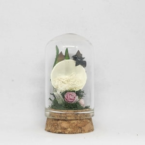 May include: A glass dome with a cork base contains a small, white, dried flower arrangement. The arrangement includes a dried flower, dried moss, and a small, black bird figurine.