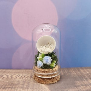 May include: A glass cloche with a cork base contains a white egg shell filled with white shredded paper and three blue flowers with green leaves.