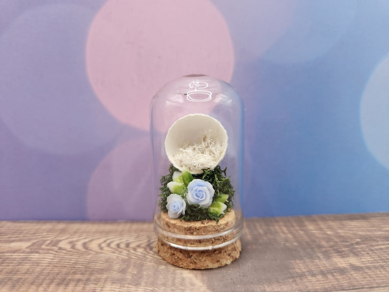 May include: A glass cloche with a cork base contains a white egg shell filled with white shredded paper and three blue flowers with green leaves.