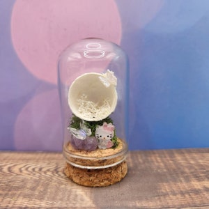 May include: A glass jar with a cork lid contains a miniature scene with a white egg shell, green moss, a purple crystal, and a small pink Hello Kitty figurine.