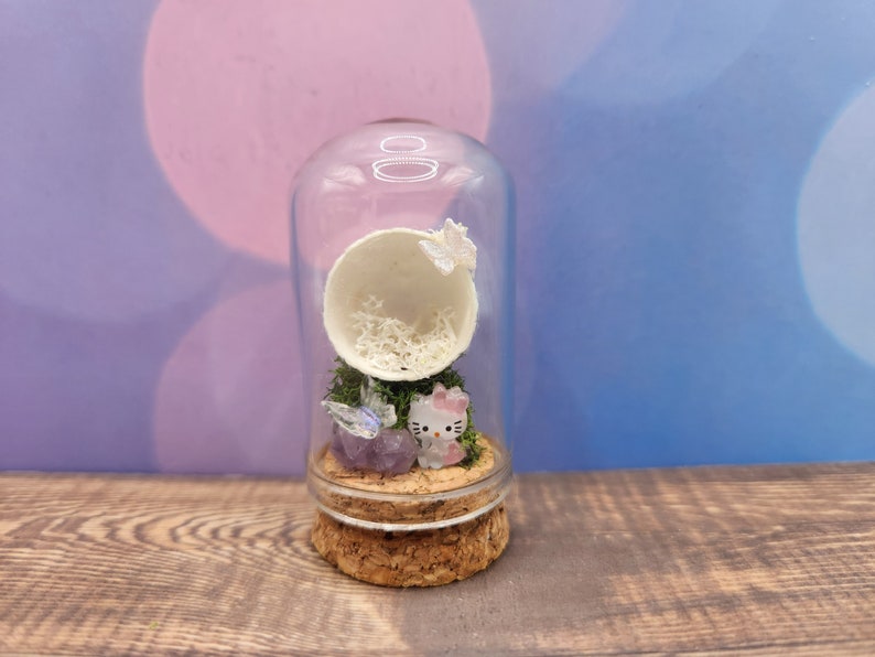 May include: A glass jar with a cork lid contains a miniature scene with a white egg shell, green moss, a purple crystal, and a small pink Hello Kitty figurine.