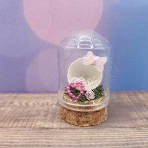 May include: A glass cloche with a cork base contains a white eggshell, pink heart-shaped beads, a pink flower, and a pink butterfly.