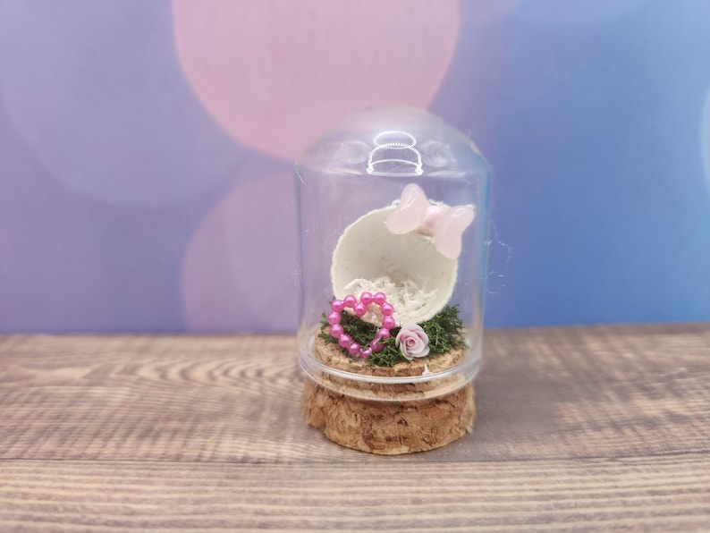 May include: A glass cloche with a cork base contains a white eggshell, pink heart-shaped beads, a pink flower, and a pink butterfly.