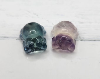May include: Two small carved fluorite crystal figurines in the shape of frogs. The frog on the left is a light blue color, while the frog on the right is a light purple color.