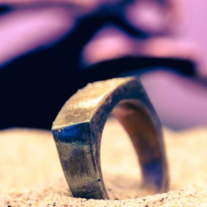 Peut inclure: Gros plan sur une bague dorée au design géométrique. La bague est partiellement enfouie dans le sable, avec un arrière-plan flou. La bague a un dessus carré et un anneau arrondi.