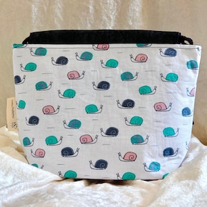 May include: A white fabric bag with a repeating pattern of colorful snails in teal, pink, and navy blue. The bag has a black top and a small tag with text. The bag is sitting on a white, textured surface.