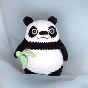 Puede incluir: Un peluche de panda tejido a crochet con pelaje blanco y negro, sosteniendo un brote de bambú verde. El panda tiene grandes ojos negros y una expresión amigable.