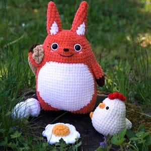 May include: A crocheted red fox plush toy holding a brown crocheted object, standing on a tree stump with a white crocheted chicken and two white crocheted eggs beside it.