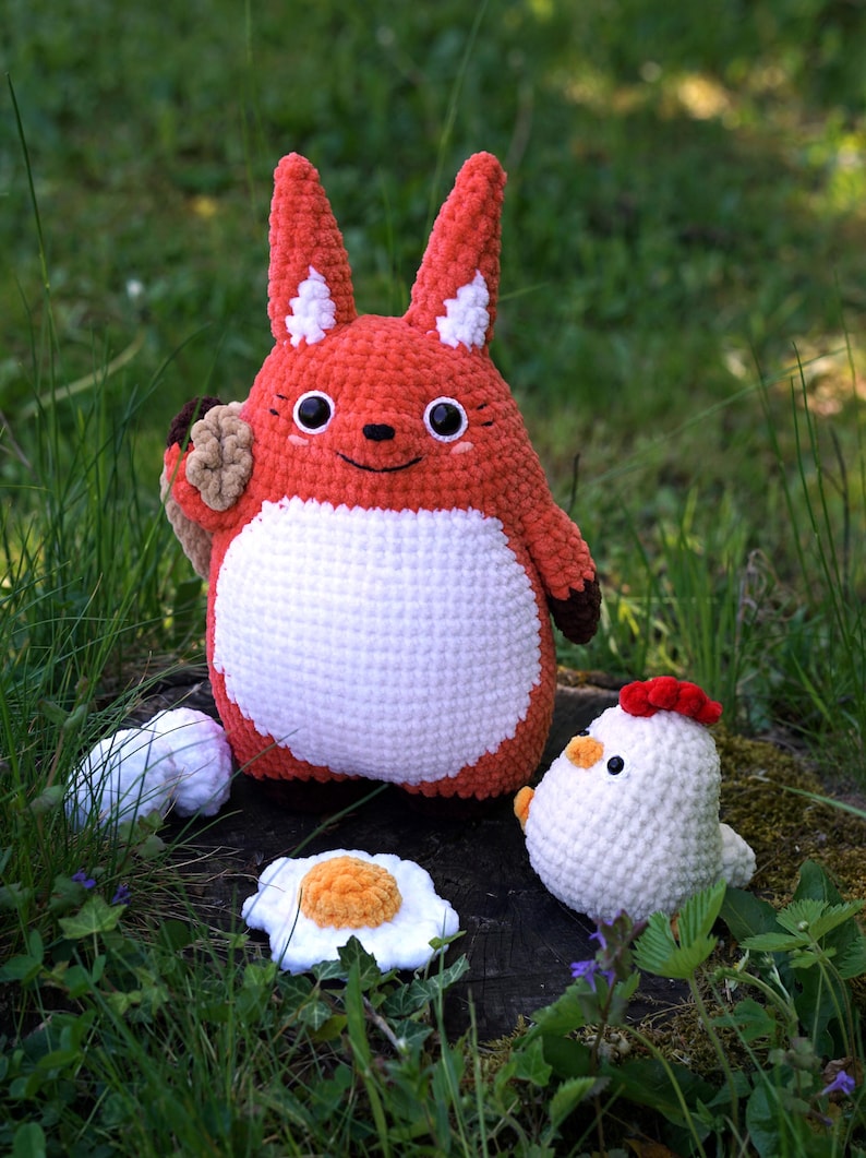 May include: A crocheted red fox plush toy holding a brown crocheted object, standing on a tree stump with a white crocheted chicken and two white crocheted eggs beside it.