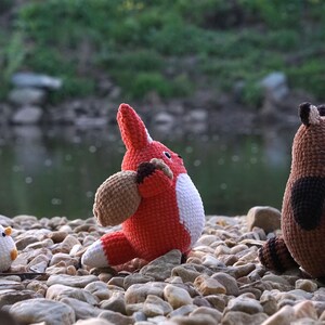 May include: Three crocheted stuffed animals: a red fox, a brown raccoon, and a white chicken. The animals are sitting on a bed of small rocks.
