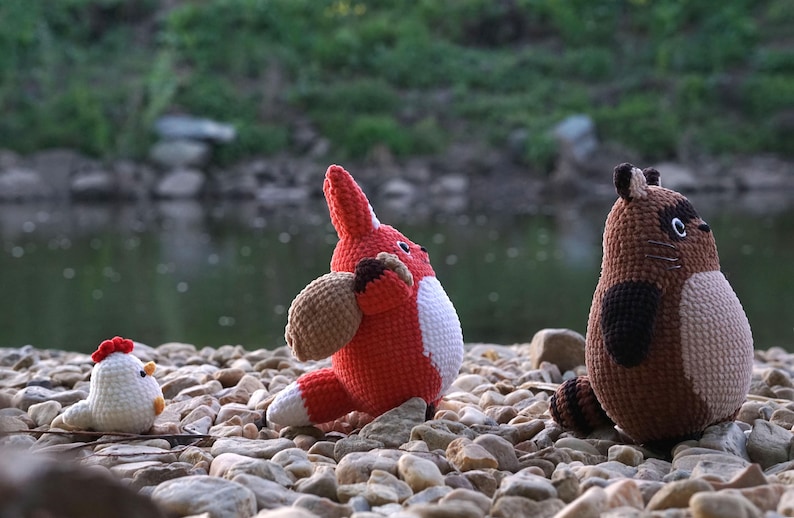May include: Three crocheted stuffed animals: a red fox, a brown raccoon, and a white chicken. The animals are sitting on a bed of small rocks.