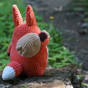 May include: A crocheted fox plush toy with a brown and white tail, sitting on a tree stump in a forest setting.
