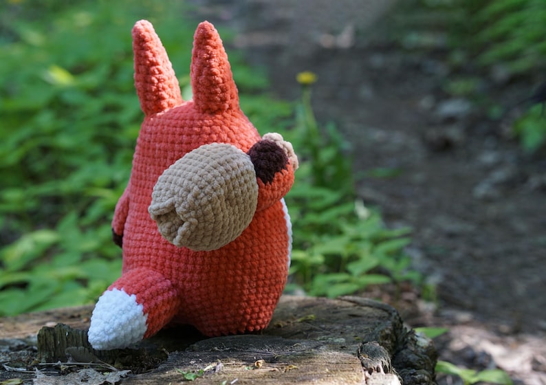 May include: A crocheted fox plush toy with a brown and white tail, sitting on a tree stump in a forest setting.