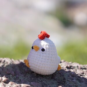 May include: A white crocheted chicken toy with a red comb and orange beak. The toy is sitting on a rocky surface.