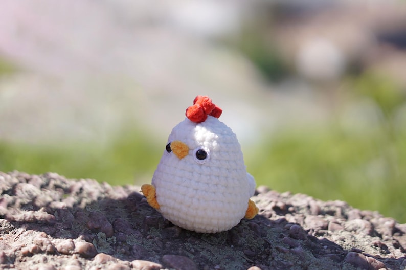 May include: A white crocheted chicken toy with a red comb and orange beak. The toy is sitting on a rocky surface.