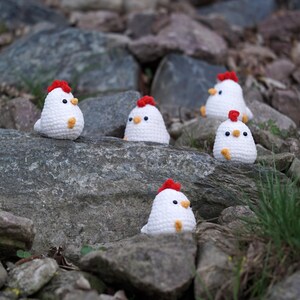 May include: Five white crocheted chicks with red combs and yellow beaks are sitting on gray rocks. The chicks are arranged in a line, with the first chick on the left and the last chick on the right.