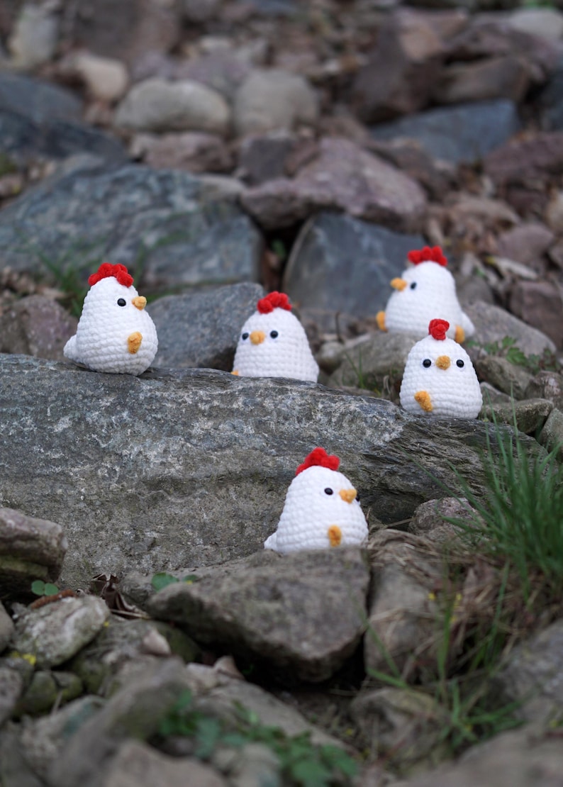 May include: Five white crocheted chicks with red combs and yellow beaks are sitting on gray rocks. The chicks are arranged in a line, with the first chick on the left and the last chick on the right.