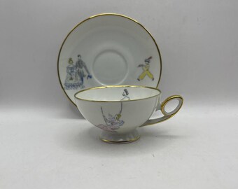 May include: White teacup and saucer set with gold trim and whimsical illustrations. The teacup features a figure on a swing, while the saucer displays figures in period clothing. A charming addition to any tea set collection.