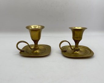 May include: Two gold-toned brass candle holders with a small square base and a handle. Each holder has a small cup for a candle.