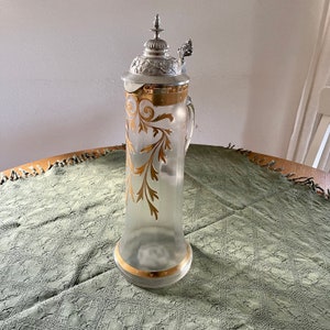 May include: A clear glass pitcher with a silver lid and a gold floral design. The pitcher has a handle and a round base.