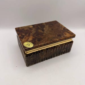 Könnte beinhalten: Eine rechteckige Schatulle aus braunem, marmoriertem Stein mit einer texturierten Basis mit vertikalen Rillen und einem glatten Deckel. Ein goldfarbener Metallrand trennt den Deckel von der Basis. Ein ovaler Aufkleber auf dem Deckel trägt die Aufschrift "Vero Alabastro Ali Lavorato a Volterra".