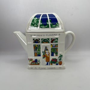 Puede incluir: Una tetera de cerámica blanca con forma de tienda de flores con un techo verde, azul y blanco. La tetera tiene un diseño floral y el texto "Florie's Flowers" y "a floral teapot".