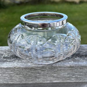 Könnte beinhalten: Eine klare, runde Glasschale mit komplexen Schliffmustern auf der Außenfläche. Die Schale ist mit einem glatten, polierten silberfarbenen Metallrand versehen, der die Öffnung umschließt und einen Kontrast zum strukturierten Glaskörper bildet.