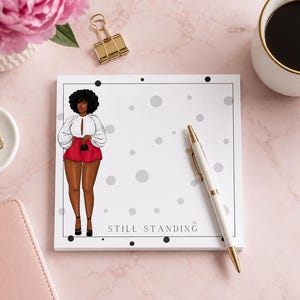 May include: A white notepad featuring an illustration of a woman in a white top and red shorts, with the text "STILL STANDING". A white and gold pen lies on the pad, alongside a coffee cup and stationery on a pink surface.