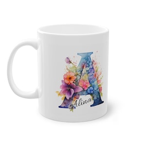 Op de afbeelding: Witte keramische mok met een aquarelontwerp van de blauwe letter "A", versierd met kleurrijke bloemen. De naam "Alina" staat onder de letter in een scriptlettertype. De mok heeft een gebogen handvat.