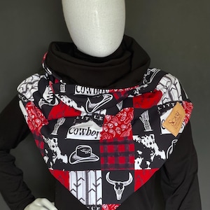 May include: A black and red triangular scarf with a patchwork design featuring cowboys, hats, and other western motifs. The scarf has a white background with black and red accents. The text "Little Cowboy" is printed on the scarf.