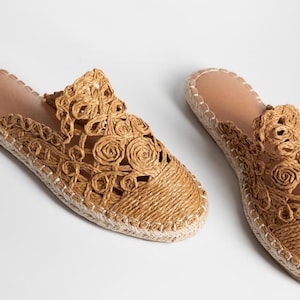 May include: A pair of espadrille slip-on shoes in a natural tan colour. The uppers feature a decorative floral design. The soles are made of woven rope, stitched with white thread. The insoles are a light brown colour.