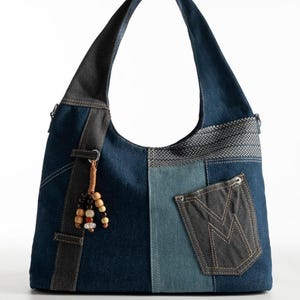 Pode incluir: Bolsa de ombro patchwork de ganga com uma única alça. A bolsa apresenta uma variedade de tons de ganga azul, um bolso com costuras decorativas e um berloque de contas. A alça e os detalhes são em ganga mais escura.
