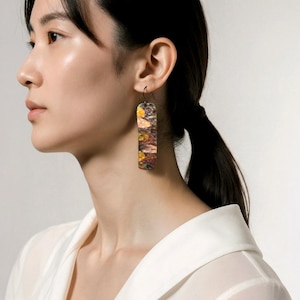 Może przedstawiać: Prostokątne kolczyki wiszące z kolorowym, abstrakcyjnym wzorem. Kolczyki mają mieszankę żółtych, brązowych i pomarańczowych odcieni. Kolczyki są zawieszone na czarnym haczyku. Nosi je osoba z ciemnymi włosami.