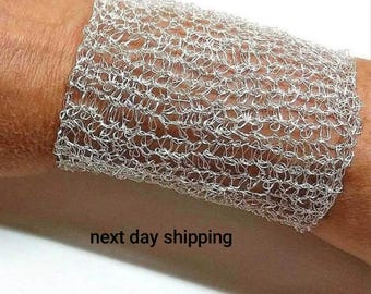 Può includere: Un bracciale largo a fascia realizzato in filo metallico color argento, caratterizzato da un intricato motivo a rete traforata che avvolge il polso. Il materiale metallico ha una lucentezza riflettente, creando un aspetto testurizzato e arioso.