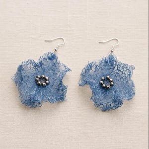Pu&ograve; includere: Un paio di orecchini a forma di fiore blu realizzati in una sottile e delicata rete metallica. Ogni orecchino presenta un gruppo centrale di piccole perline metalliche scure disposte a cerchio, sospese a un semplice gancio in metallo argentato.
