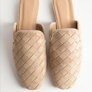 May include: A pair of beige woven slip-on mules. The shoes have a textured, basket-weave design on the upper and a smooth, neutral-toned insole. The open-back design and muted color palette suggest a casual, yet stylish aesthetic.