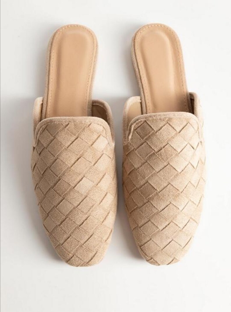 May include: A pair of beige woven slip-on mules. The shoes have a textured, basket-weave design on the upper and a smooth, neutral-toned insole. The open-back design and muted color palette suggest a casual, yet stylish aesthetic.