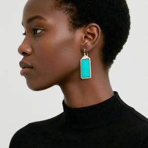 Op de afbeelding: Goudkleurige rechthoekige oorbellen met een helder turquoise middenstuk. De oorbellen hebben een eenvoudig, elegant ontwerp, met een rechthoekige turkooizen steen gezet in een gouden montuur. De oorbellen hangen aan een kleine gouden haak.