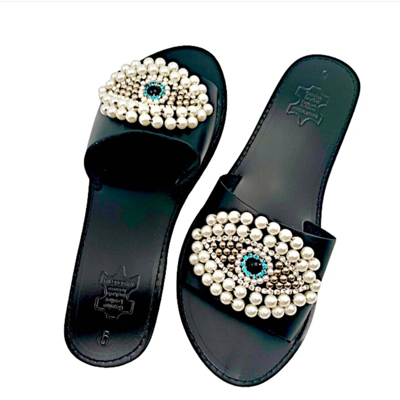 May include: Black leather slide sandals with a pearl and turquoise evil eye embellishment. The sandals have a simple design and are perfect for summer.
