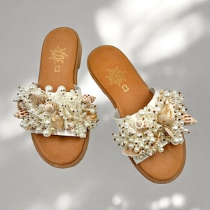 May include: A pair of white leather sandals with a brown sole. The sandals are decorated with a cluster of white pearls and seashells.