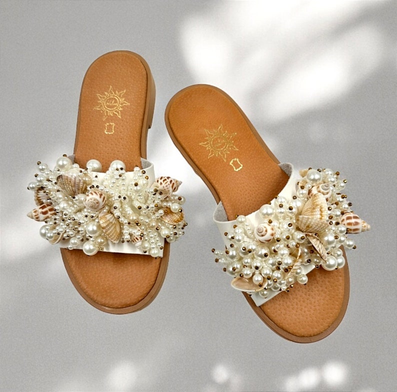 May include: A pair of white leather sandals with a brown sole. The sandals are decorated with a cluster of white pearls and seashells.