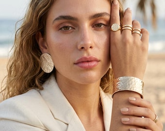 May include: A set of gold-toned jewelry featuring a large circular hammered earring, a wide hammered cuff bracelet, and three stacked rings on the fingers. The jewelry has a consistent textured finish and a polished metallic appearance.