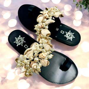 Könnte beinhalten: Schwarze Ledersandalen mit einem großen Schmuckstück aus Muscheln und Perlen. Die Sandalen haben ein schlichtes Design mit einem Zehenriemen und einem Riemen über dem Fußrücken.